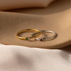 Two delicate rings—a gold Mini Toi et Moi Birthstone Ring with a green birthstone and a silver ring with a red stone—rest gracefully on a soft beige surface.