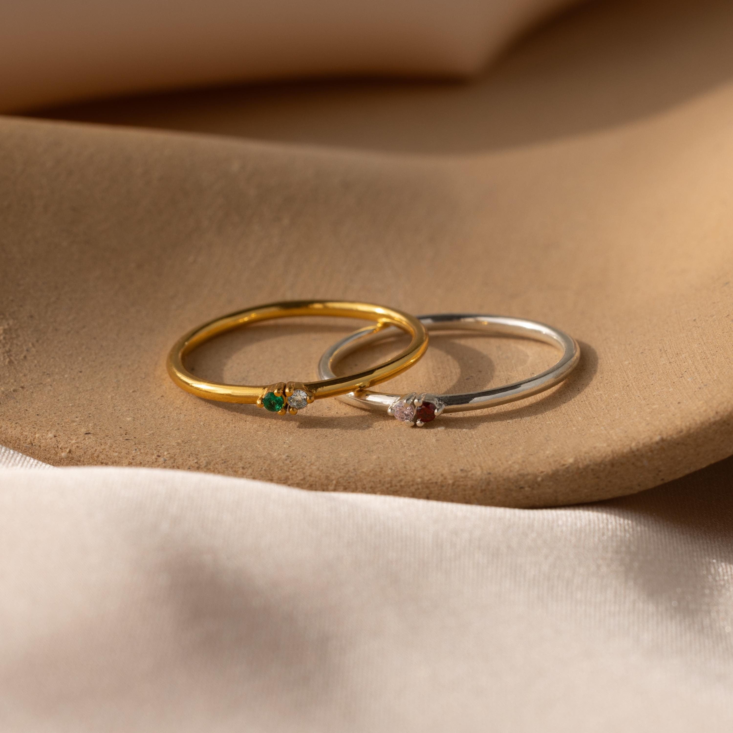 Two delicate rings—a gold Mini Toi et Moi Birthstone Ring with a green birthstone and a silver ring with a red stone—rest gracefully on a soft beige surface.