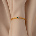 Gold bangle with green and white gemstones displayed on beige fabric—pair it with the Mini Toi et Moi Birthstone Ring for a timeless look.