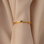 Gold bangle with green and white gemstones displayed on beige fabric—pair it with the Mini Toi et Moi Birthstone Ring for a timeless look.