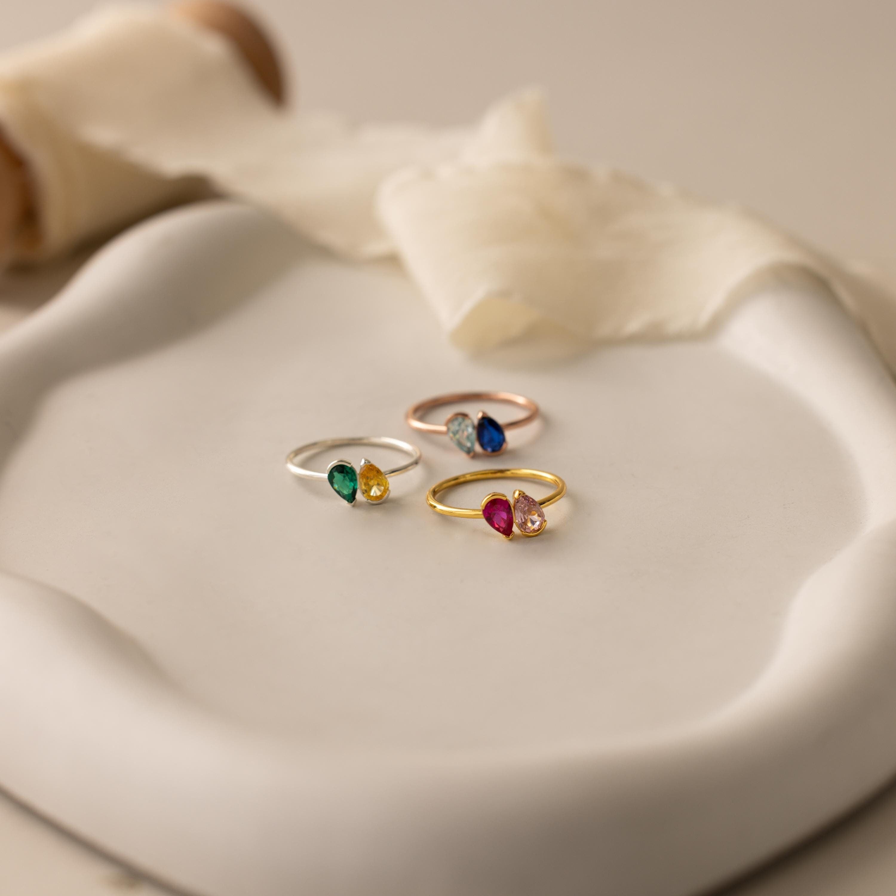 The Toi et Moi Pear Birthstone Ring, a vibrant and romantic piece, rests on a white ceramic dish with a soft cream ribbon behind it—making it an ideal gift.