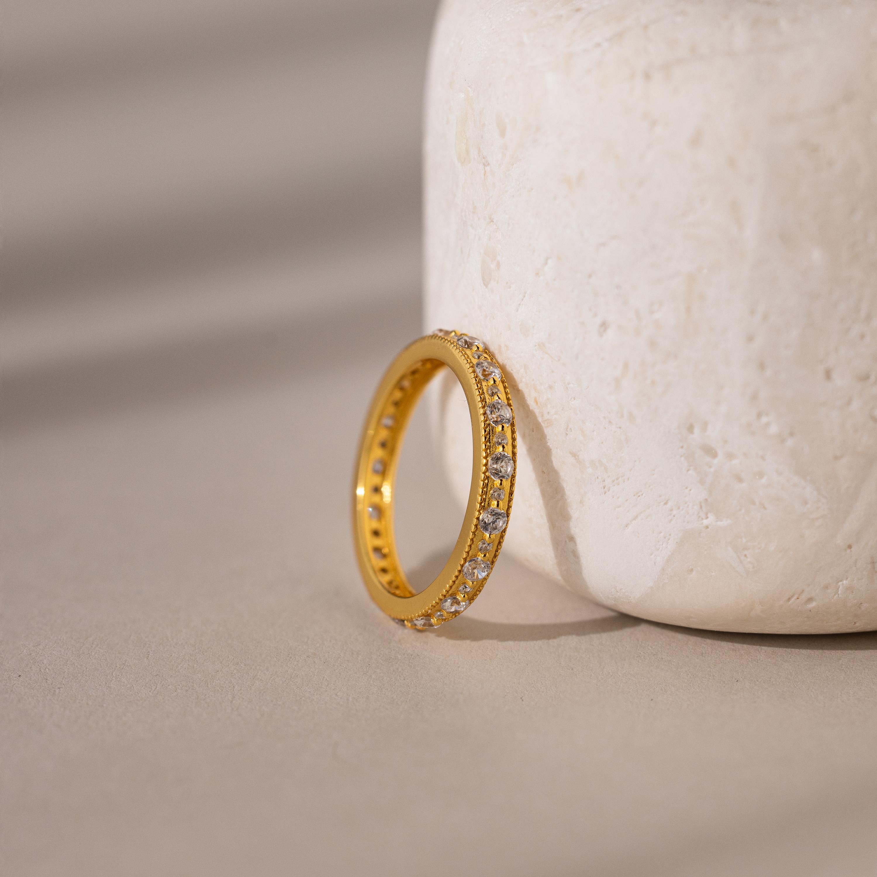 The Diamond Eternity Ring features sparkling diamonds set in gold and rests against a textured, light stone—an elegant, stackable piece ideal for any occasion.