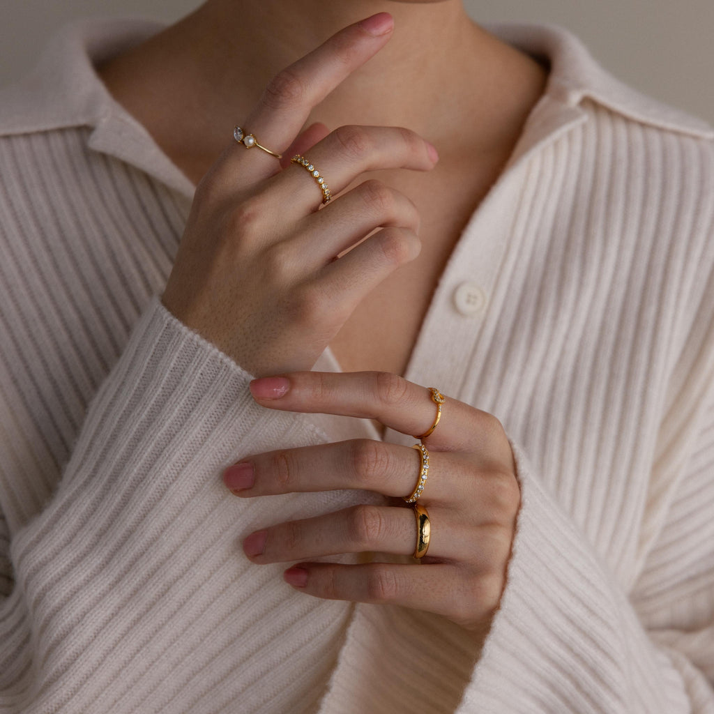 A person in a cream ribbed shirt wears several gold rings, including the Diamond Eternity Ring, stacked on their fingers with hands near their face.