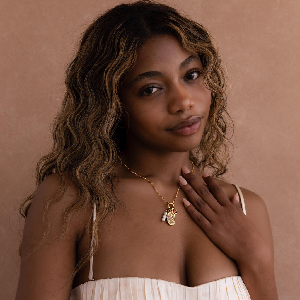 A woman with wavy hair in a cream dress touches her Love Poem Pendant Necklace, a timeless symbol of love, and looks at the camera against a beige background.