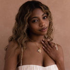 A woman with wavy hair in a cream dress touches her Love Poem Pendant Necklace, a timeless symbol of love, and looks at the camera against a beige background.