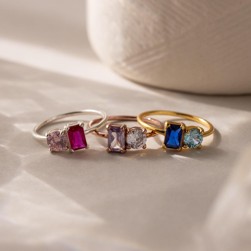 Four gemstone rings, including the Toi et Moi Emerald Birthstone Ring, are shown in various colors and metals on a sunlit surface.