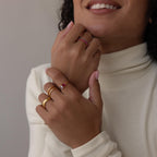 Woman in a cream turtleneck smiles and displays her hands adorned with gold rings, including a stacking birthstone ring and the elegant Open Toi et Moi Birthstone Ring.