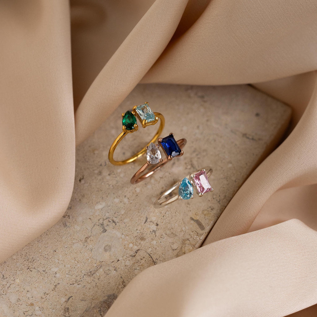 The Open Toi et Moi Birthstone Ring, a customizable colorful gemstone ring, rests on a stone surface surrounded by soft beige fabric.