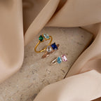 The Open Toi et Moi Birthstone Ring, a customizable colorful gemstone ring, rests on a stone surface surrounded by soft beige fabric.