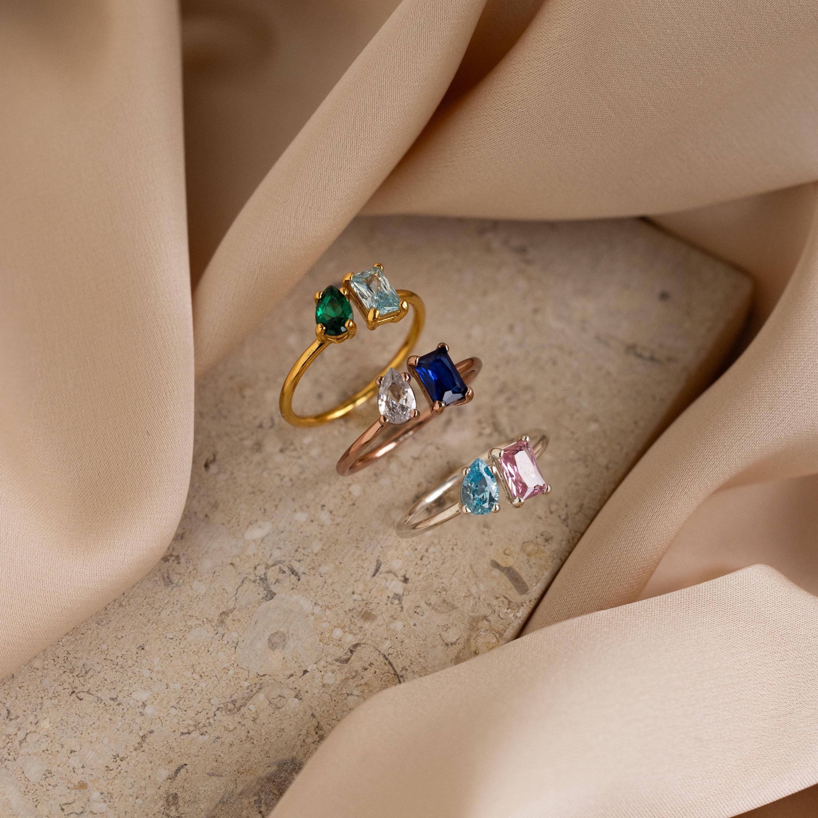 The Open Toi et Moi Birthstone Ring, a customizable colorful gemstone ring, rests on a stone surface surrounded by soft beige fabric.