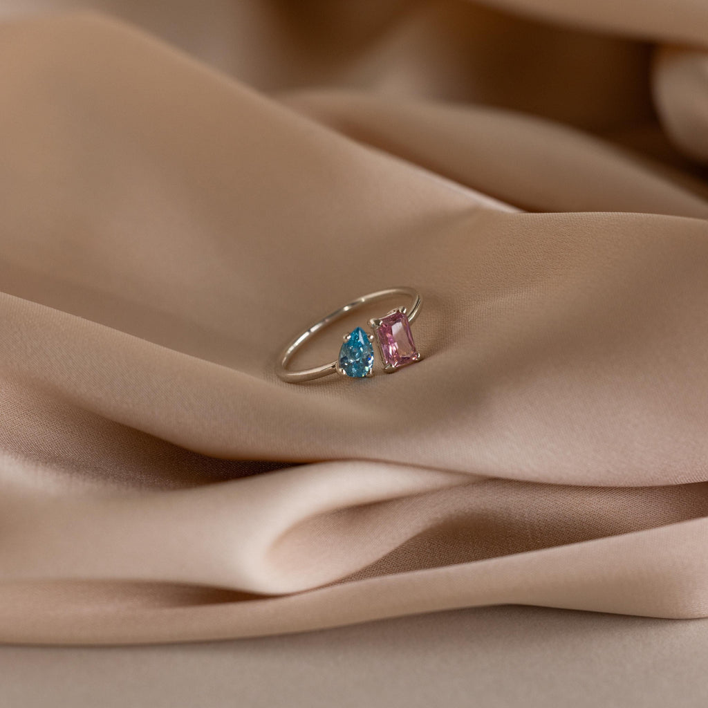 The Open Toi et Moi Birthstone Ring, featuring blue and pink gemstones, is shown resting on soft beige satin fabric.