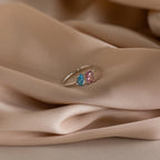The Open Toi et Moi Birthstone Ring, featuring blue and pink gemstones, is shown resting on soft beige satin fabric.