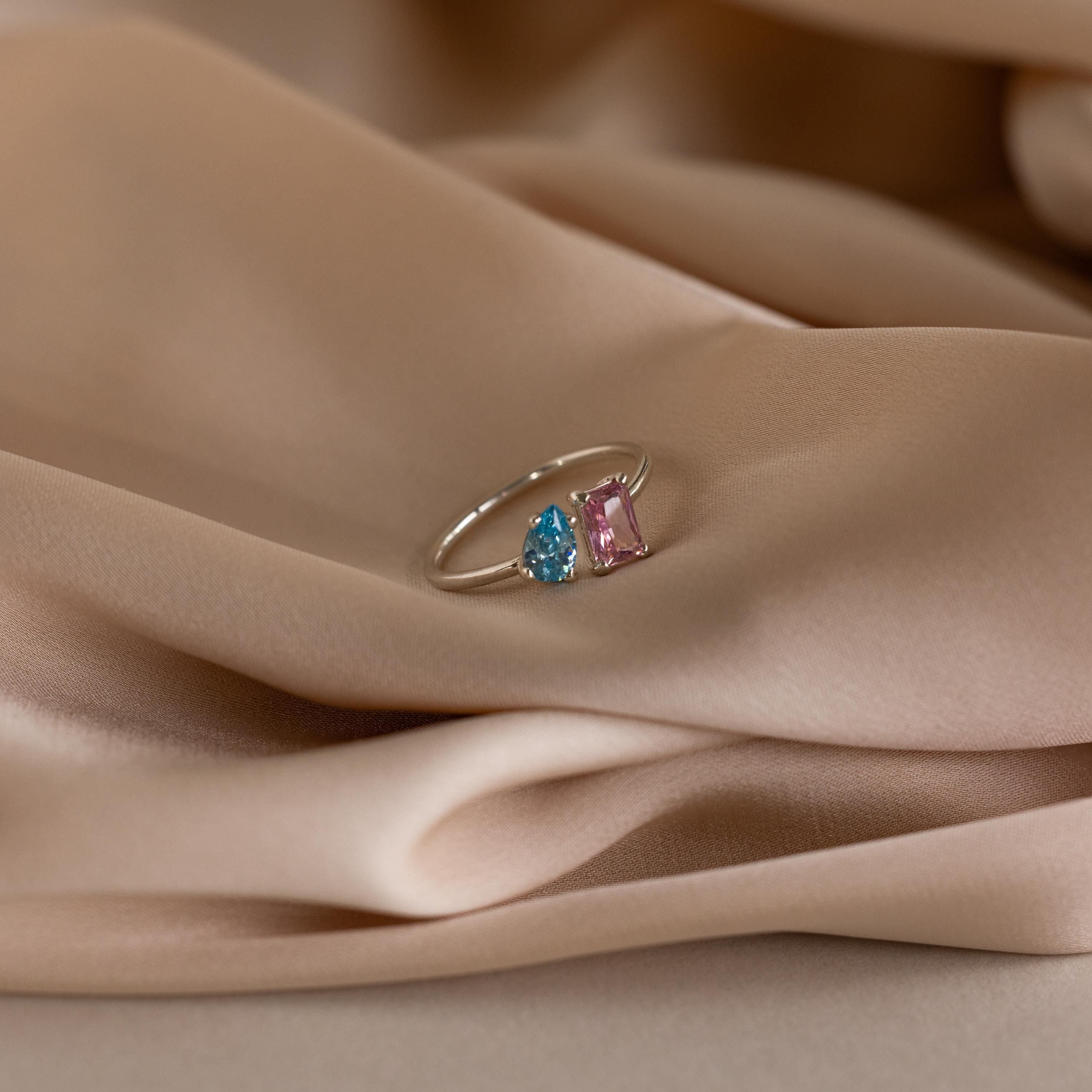 The Open Toi et Moi Birthstone Ring, featuring blue and pink gemstones, is shown resting on soft beige satin fabric.