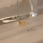 The Heart Knot Necklace, featuring a gold heart-shaped pendant on a delicate snake chain, casts a shadow on a softly lit neutral surface.