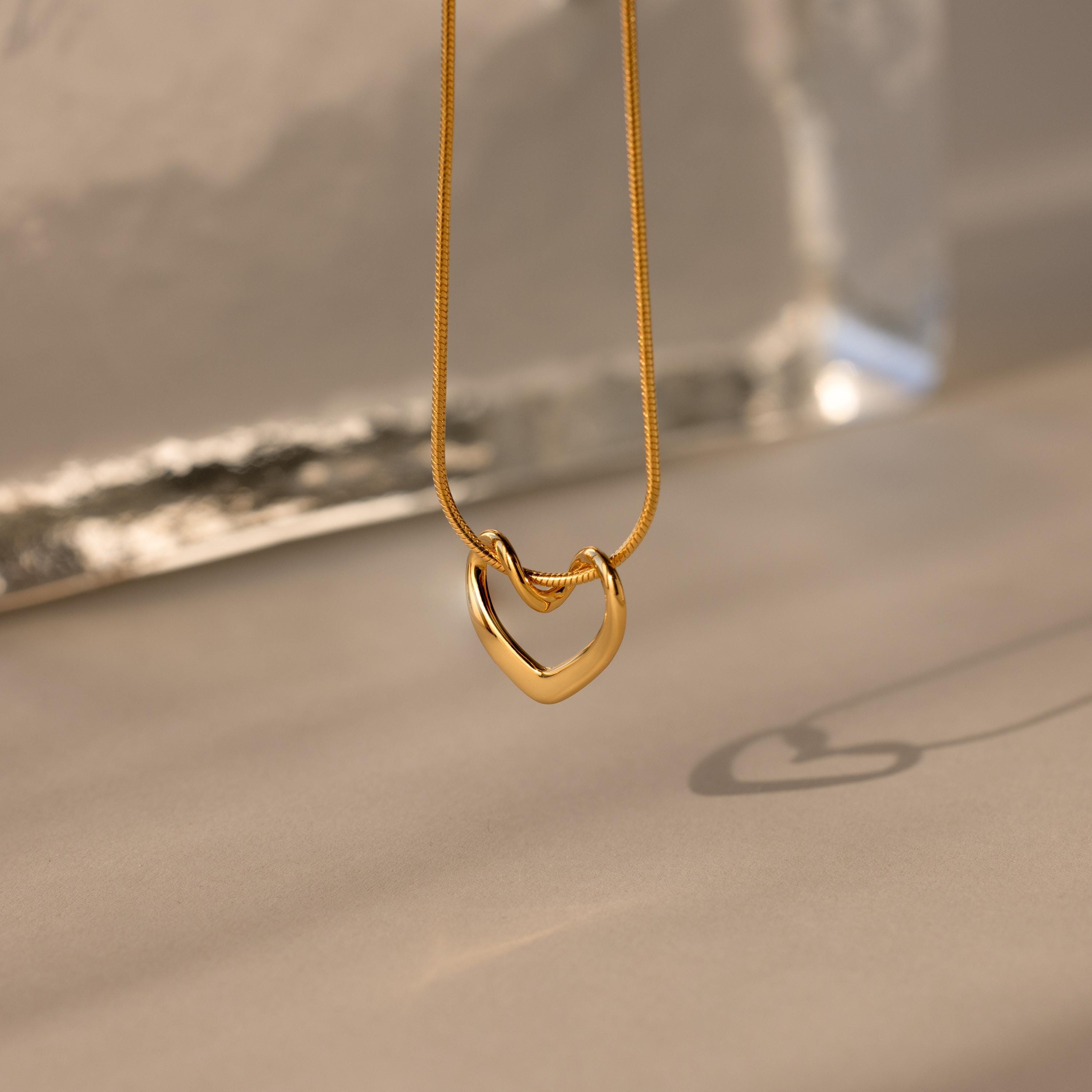 The Heart Knot Necklace, featuring a gold heart-shaped pendant on a delicate snake chain, casts a shadow on a softly lit neutral surface.