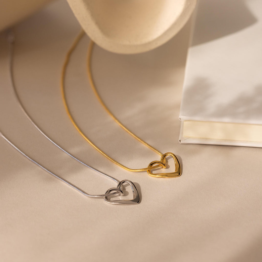 Two Heart Knot Necklaces—one gold with an open heart charm, one silver—are displayed on a beige surface next to a closed white book.