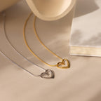 Two Heart Knot Necklaces—one gold with an open heart charm, one silver—are displayed on a beige surface next to a closed white book.