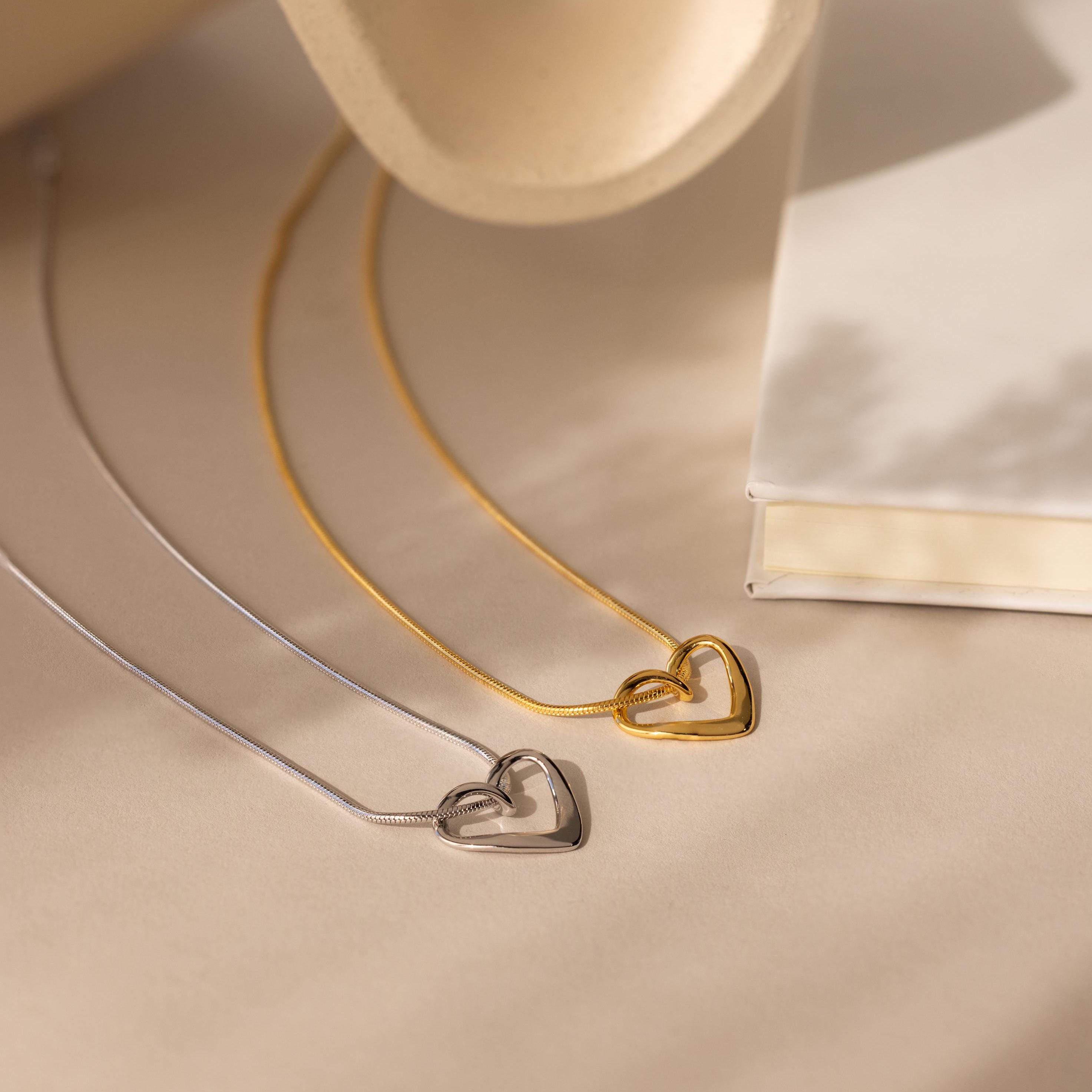 Two Heart Knot Necklaces—one gold with an open heart charm, one silver—are displayed on a beige surface next to a closed white book.