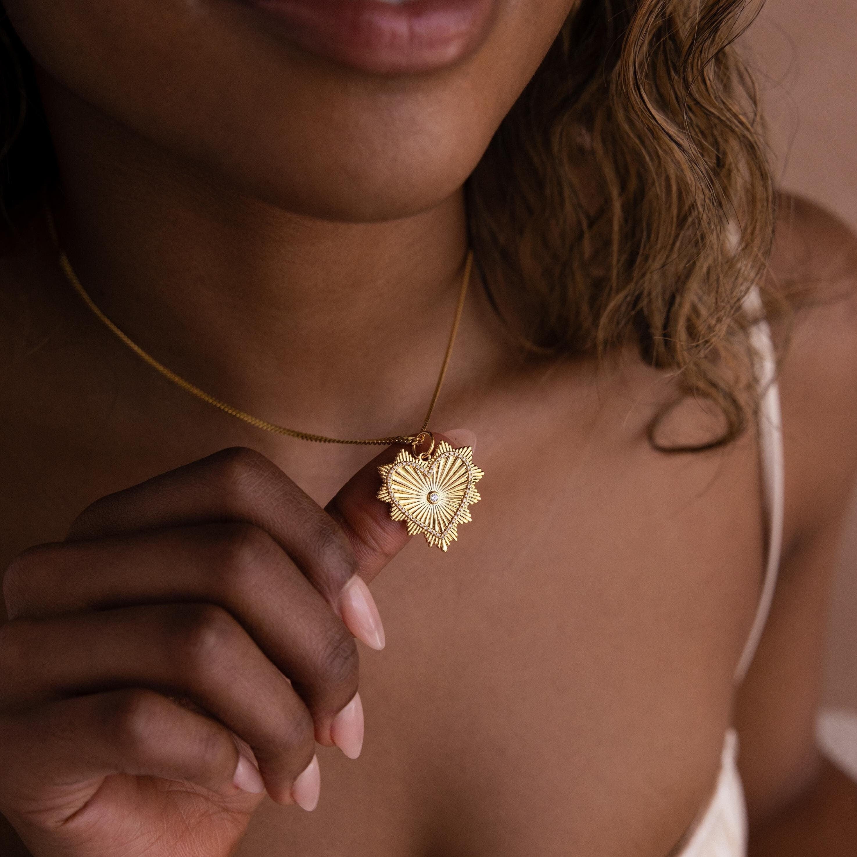 Model holds up a dainty chain with a dangling gold ribbed diamond heart charm on her neck