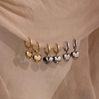 Three pairs of gold hoop earrings, including the Mixed Metal Heart Huggies and silver puff heart charms, displayed on beige fabric.