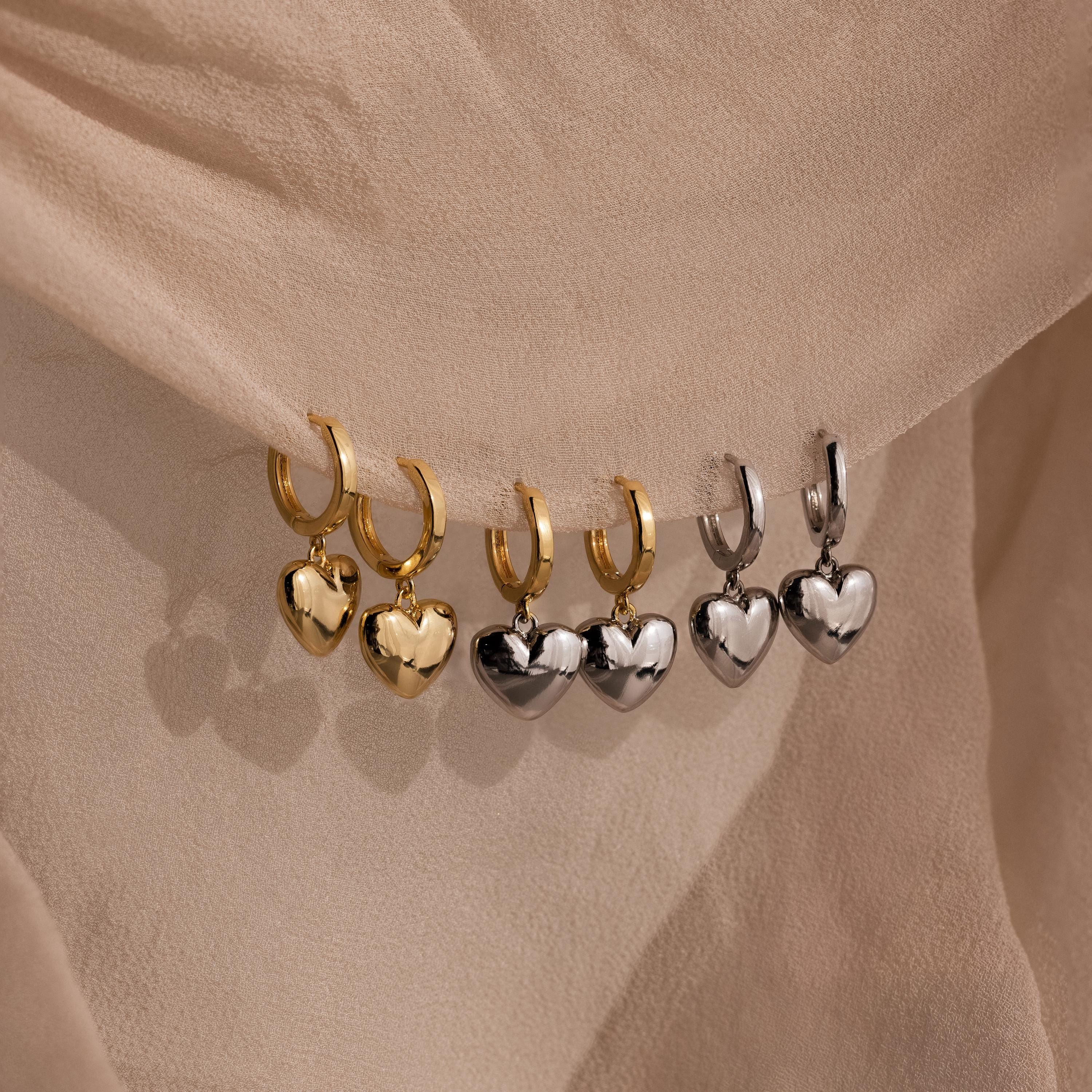 Three pairs of gold hoop earrings, including the Mixed Metal Heart Huggies and silver puff heart charms, displayed on beige fabric.