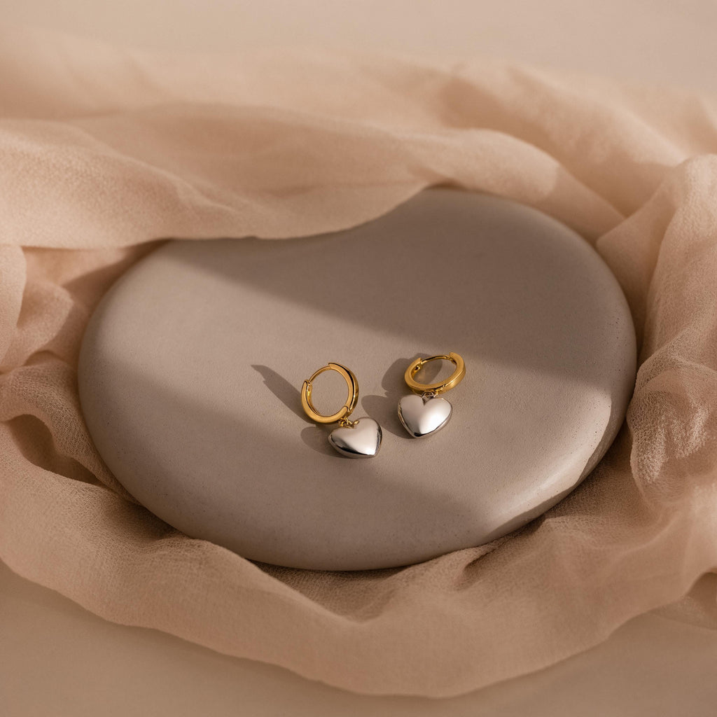 Mixed Metal Heart Huggies featuring gold hoops with silver puff heart charms, displayed on a round stone tray and accented by elegantly draped beige fabric.