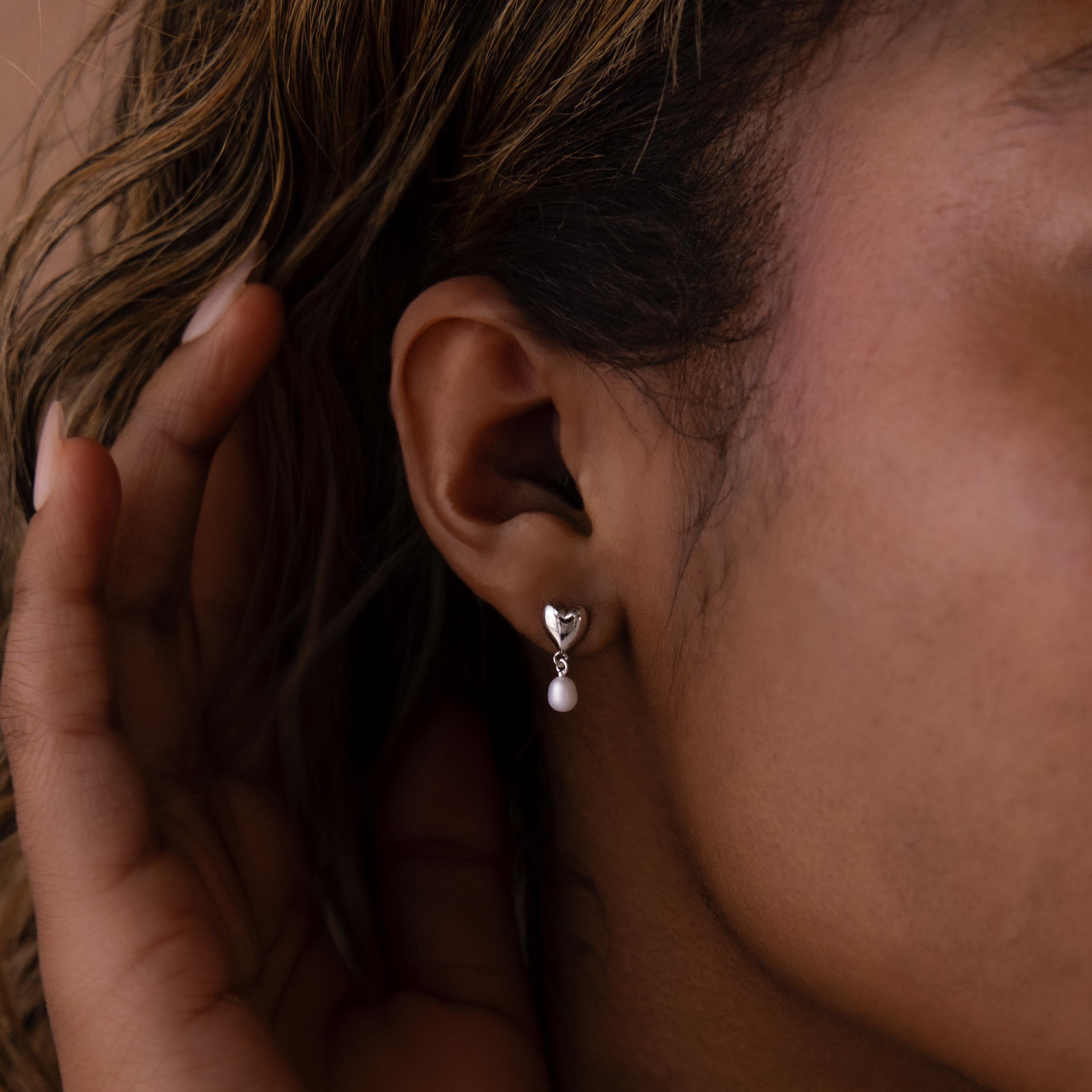 Close-up of a woman's ear wearing Heart Pearl Drop Earrings, which feature a silver heart shape and a small dangling pearl.