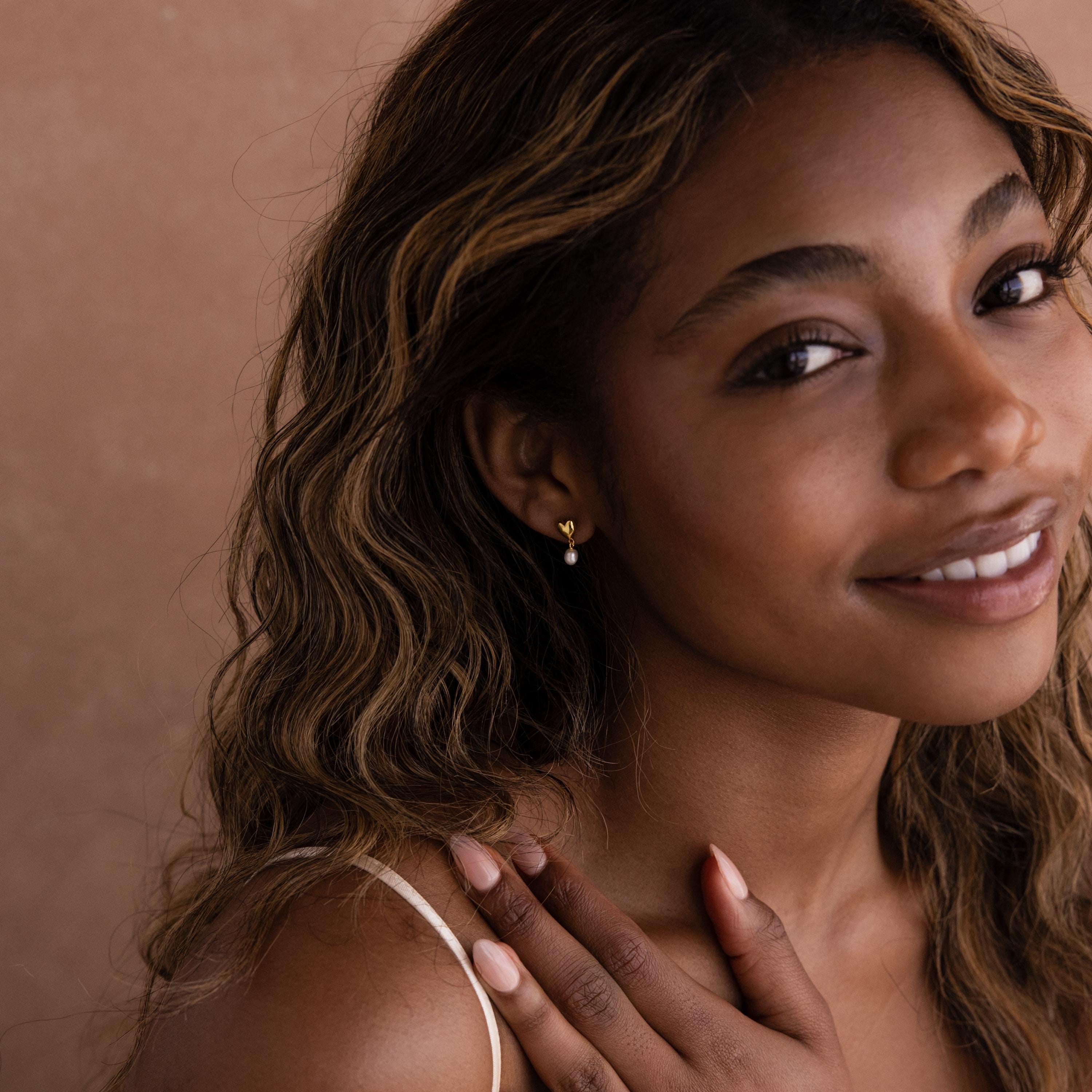 A smiling woman with wavy hair wears the elegant Heart Pearl Drop Earrings, gently touching her shoulder against a neutral backdrop.