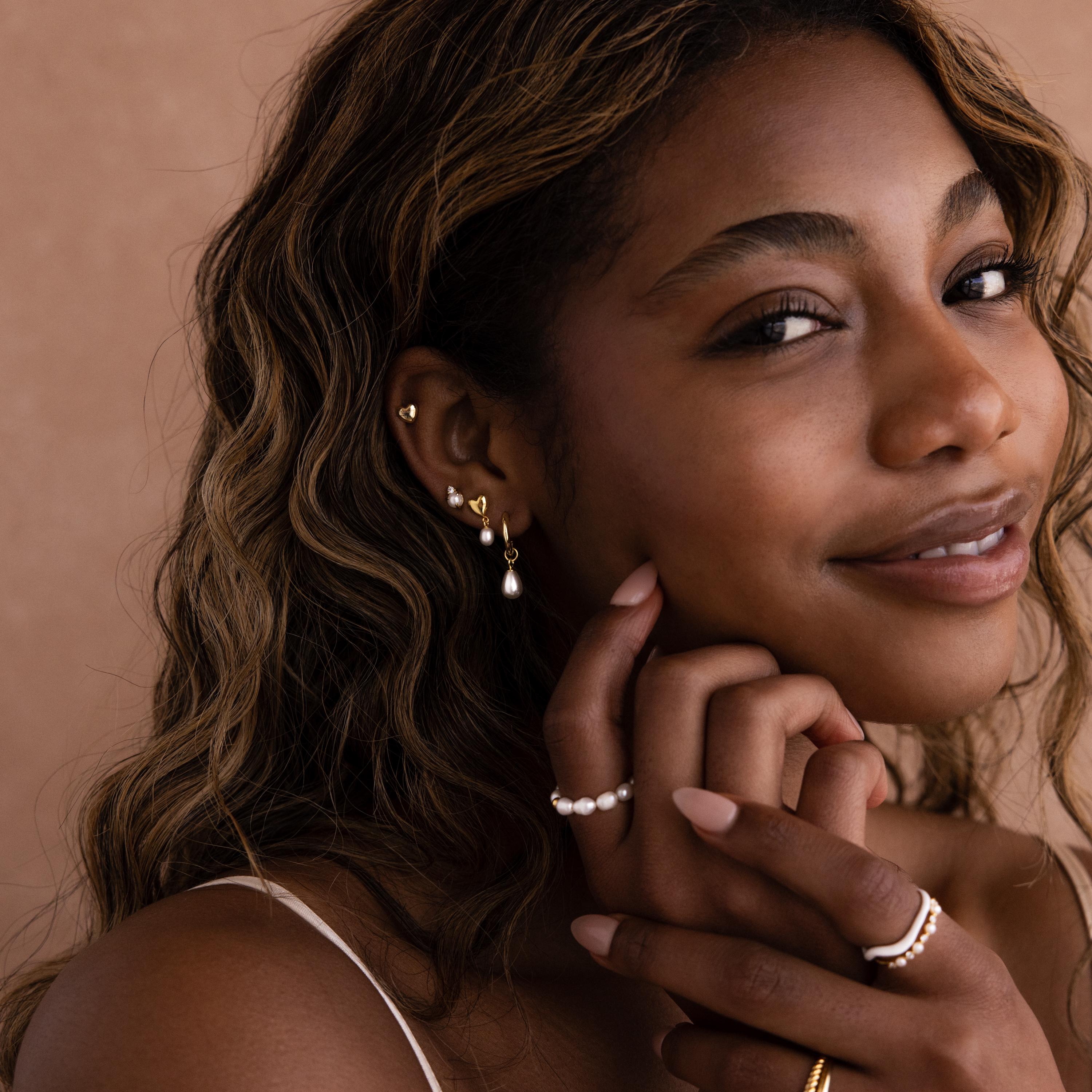 A smiling woman with wavy hair wears Heart Pearl Drop Earrings and gold rings, gently touching her face.
