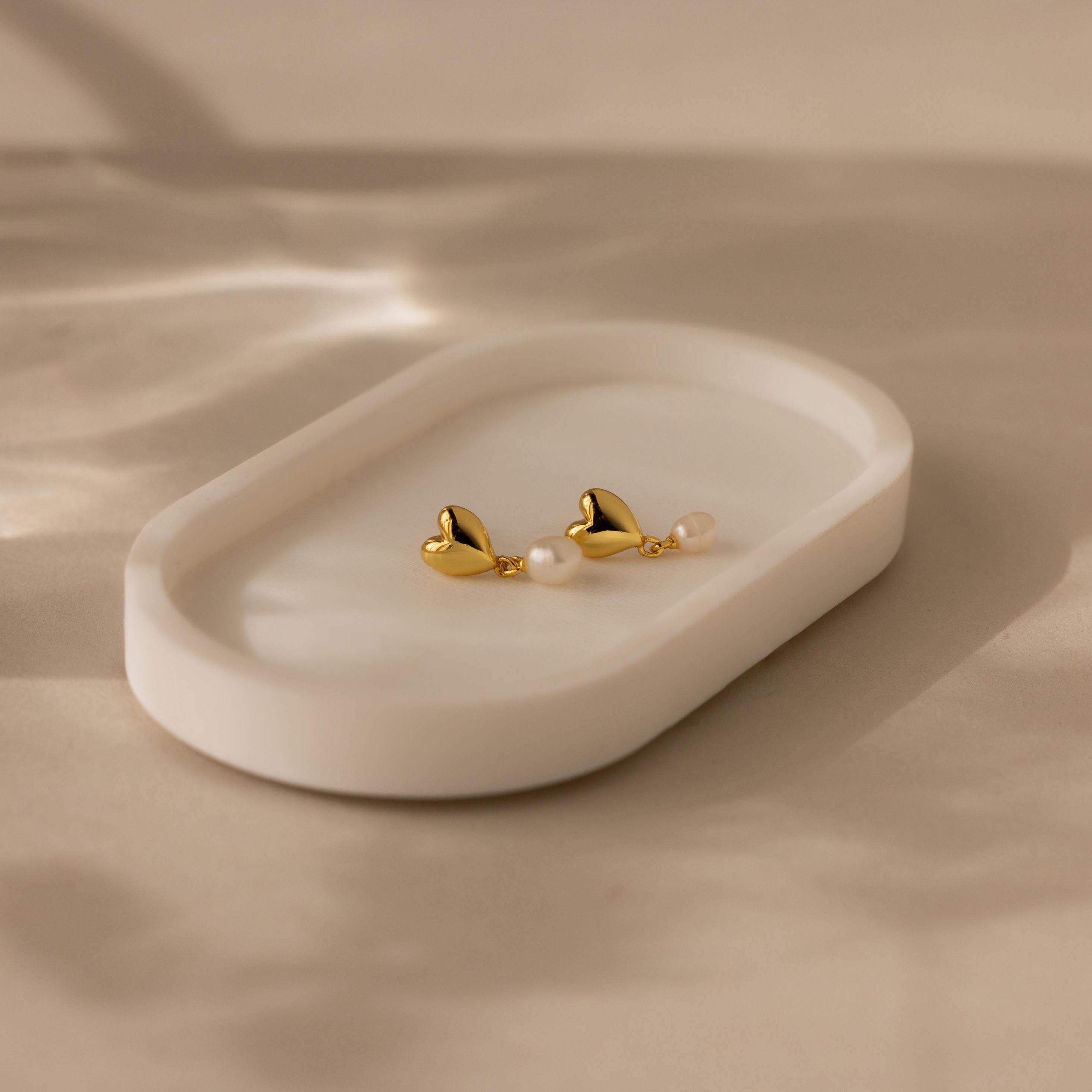 Two Heart Pearl Drop Earrings, featuring gold heart-shaped designs and delicate pearls, are displayed on a white oval ceramic dish.
