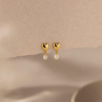 The Heart Pearl Drop Earrings, featuring gold heart studs with dangling pearls, are shown against a soft beige textured background.