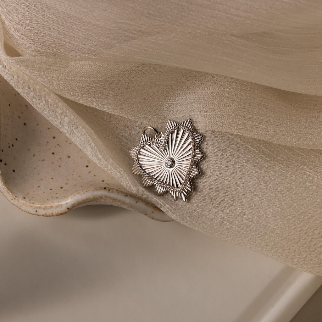 White gold ribbed heart charm with a central diamond and sunburst detailing, resting on soft fabric