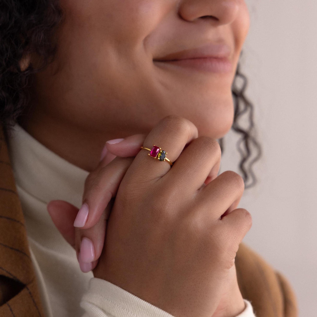 A smiling woman clasps her hands, showcasing the Toi et Moi Oval Birthstone Ring with red and green stones on her finger.