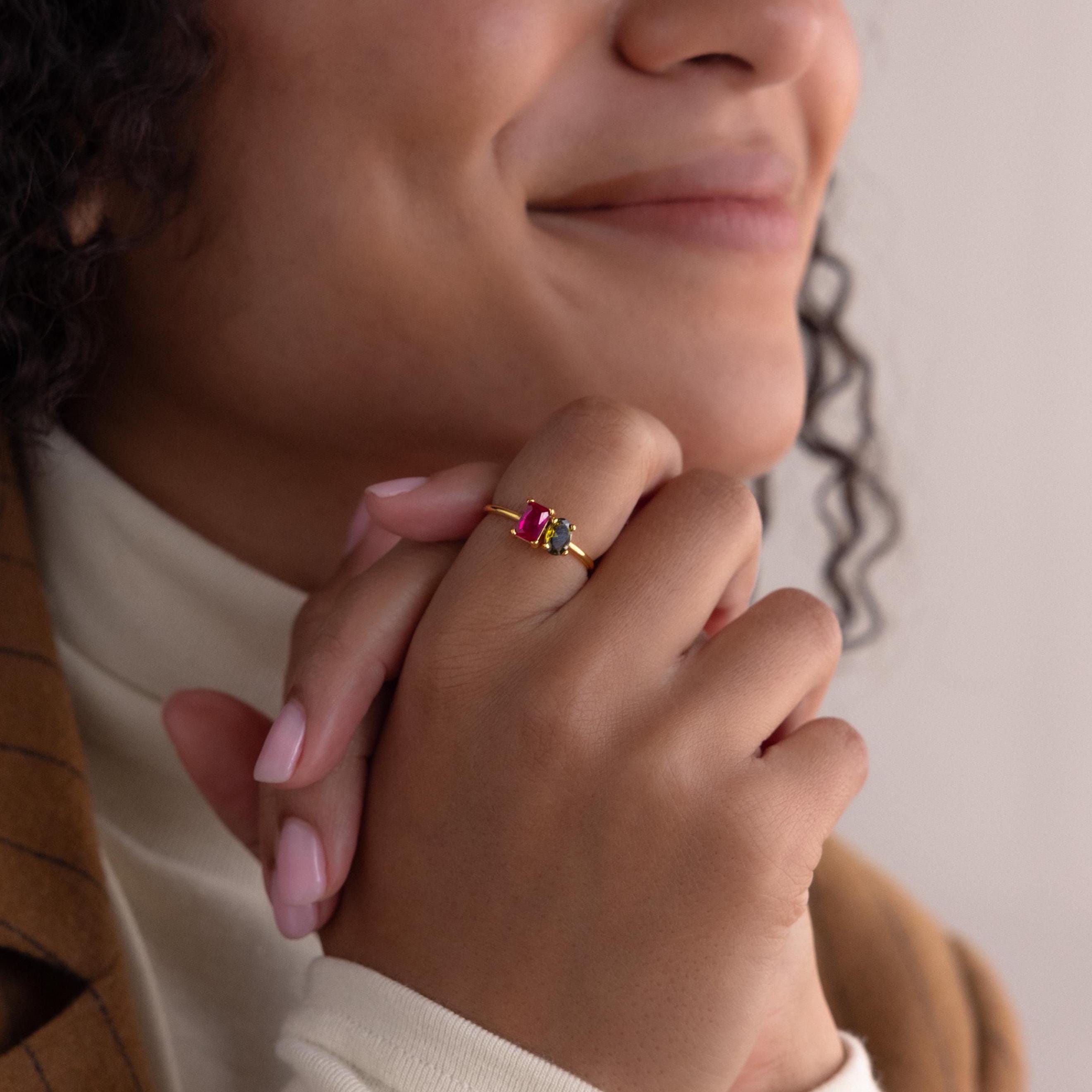 A smiling woman clasps her hands, showcasing the Toi et Moi Oval Birthstone Ring with red and green stones on her finger.