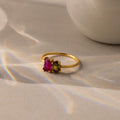 The Toi et Moi Oval Birthstone Ring, featuring one red and one dark gemstone, rests on a beige surface bathed in soft rainbow light.