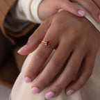 A hand with light pink nails wears a gold Toi et Moi Oval Birthstone Ring featuring two oval gemstones, one red and one green.