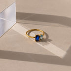 The Toi et Moi Oval Birthstone Ring, with blue and light blue gemstones, rests on a beige surface in sunlight near a clear acrylic block.