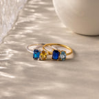 Two elegant Toi et Moi Oval Birthstone Rings with blue and yellow gemstones rest on a textured surface beside a white object.