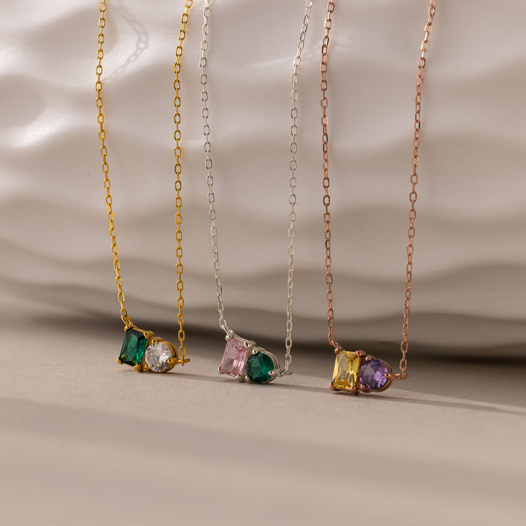 The Toi et Moi Emerald Birthstone Necklace, one of three delicate chain necklaces, features a colorful gemstone pendant displayed against a textured white background.