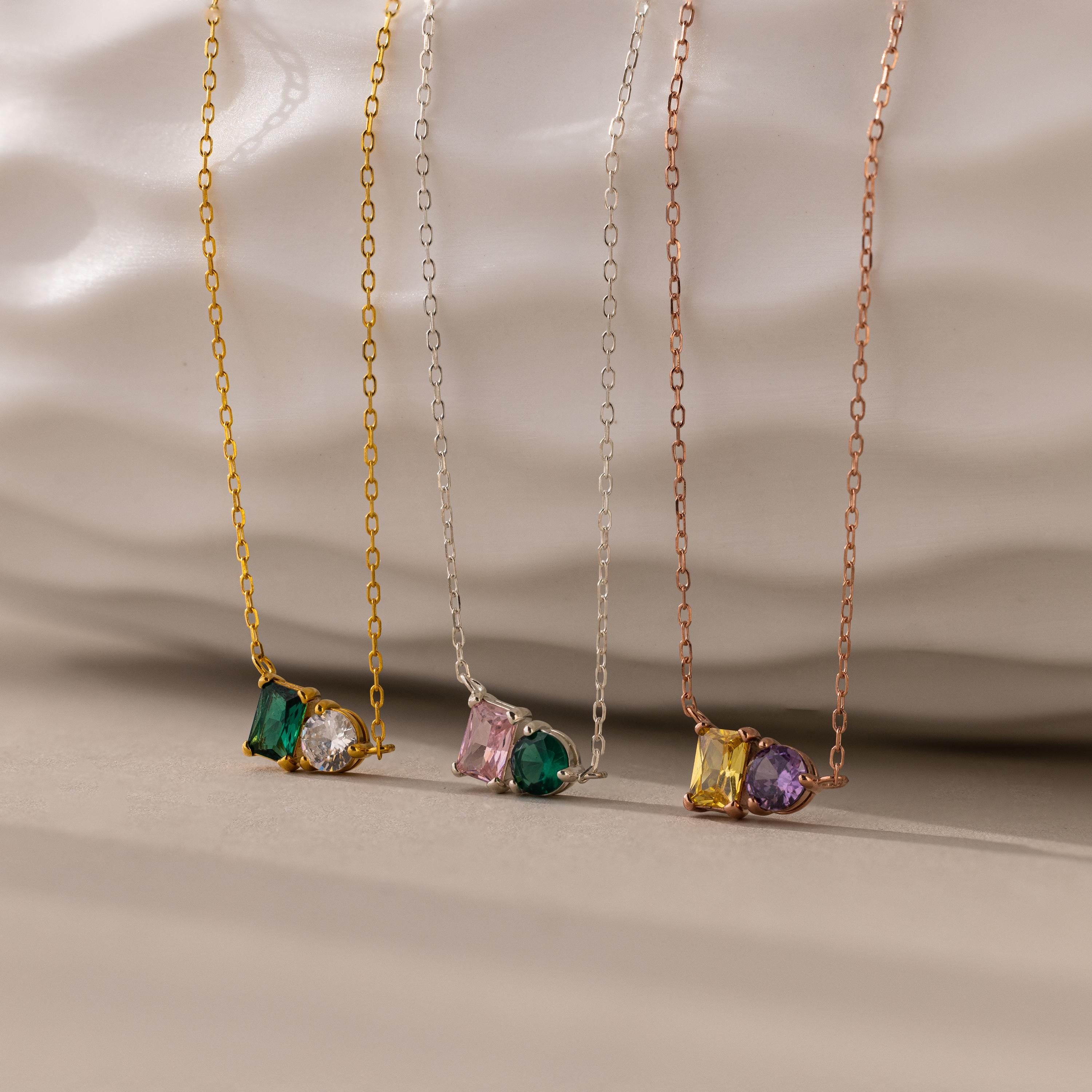 The Toi et Moi Emerald Birthstone Necklace, one of three delicate chain necklaces, features a colorful gemstone pendant displayed against a textured white background.