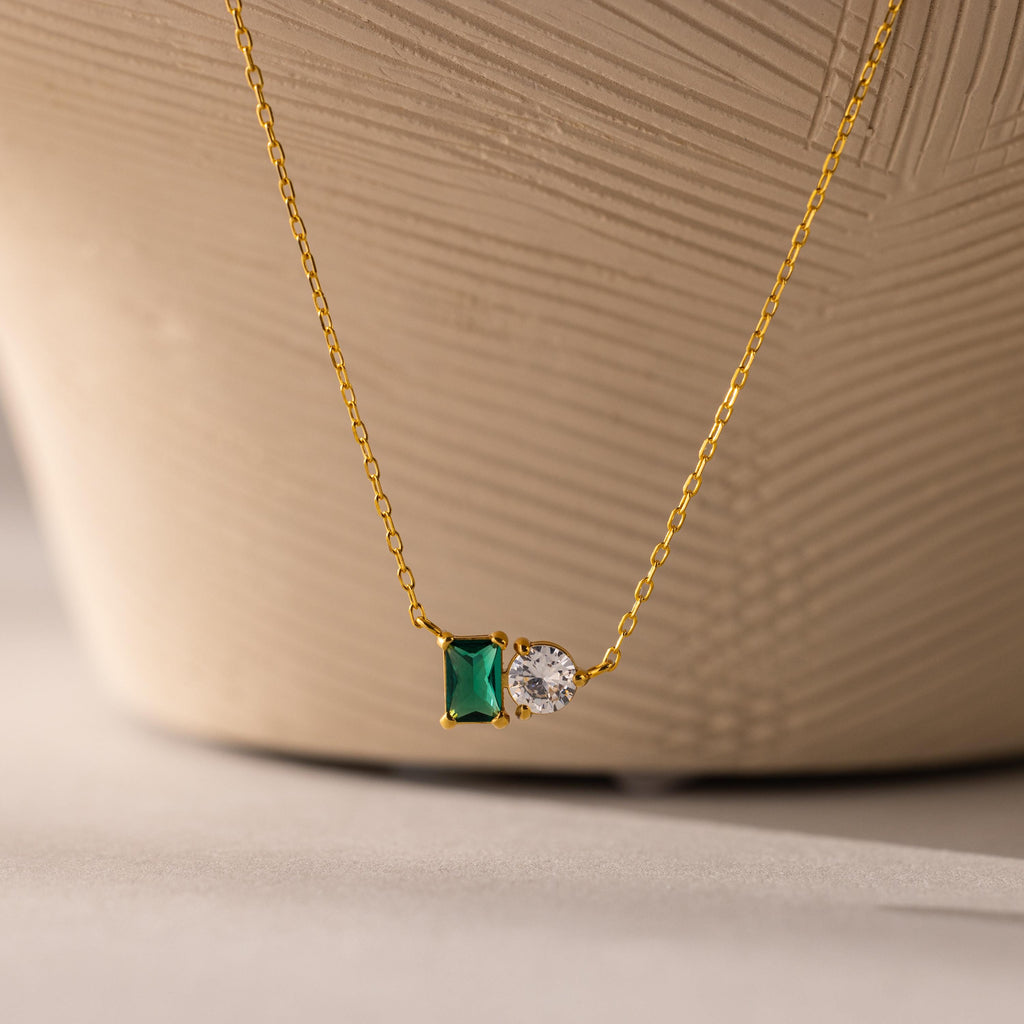 The Toi et Moi Emerald Birthstone Necklace features a green rectangular gemstone and a round white stone pendant, showcased against a textured background.