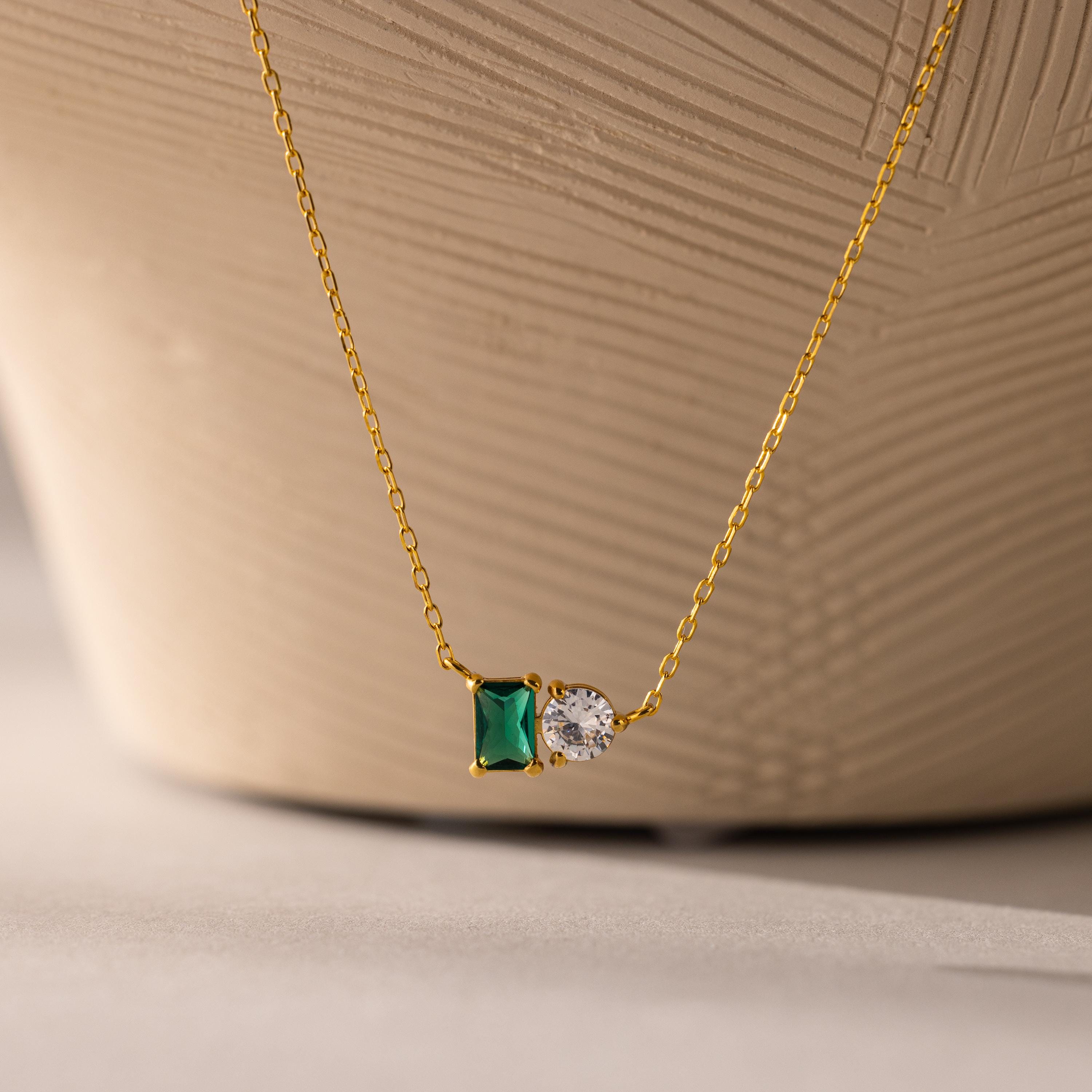 The Toi et Moi Emerald Birthstone Necklace features a green rectangular gemstone and a round white stone pendant, showcased against a textured background.