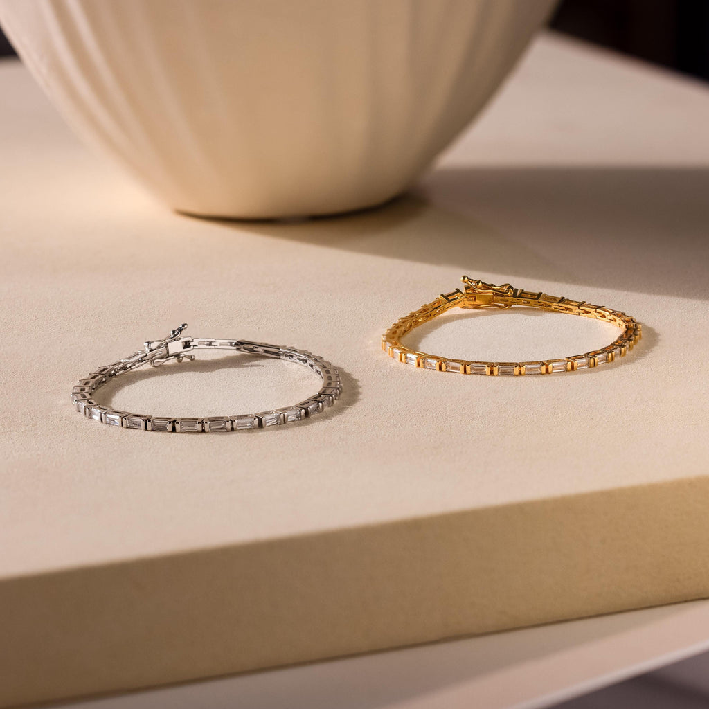 Two Claudia Baguette Tennis Bracelets, in silver and gold, shine on a light beige surface by a vase—an elegant showcase of timeless jewelry.