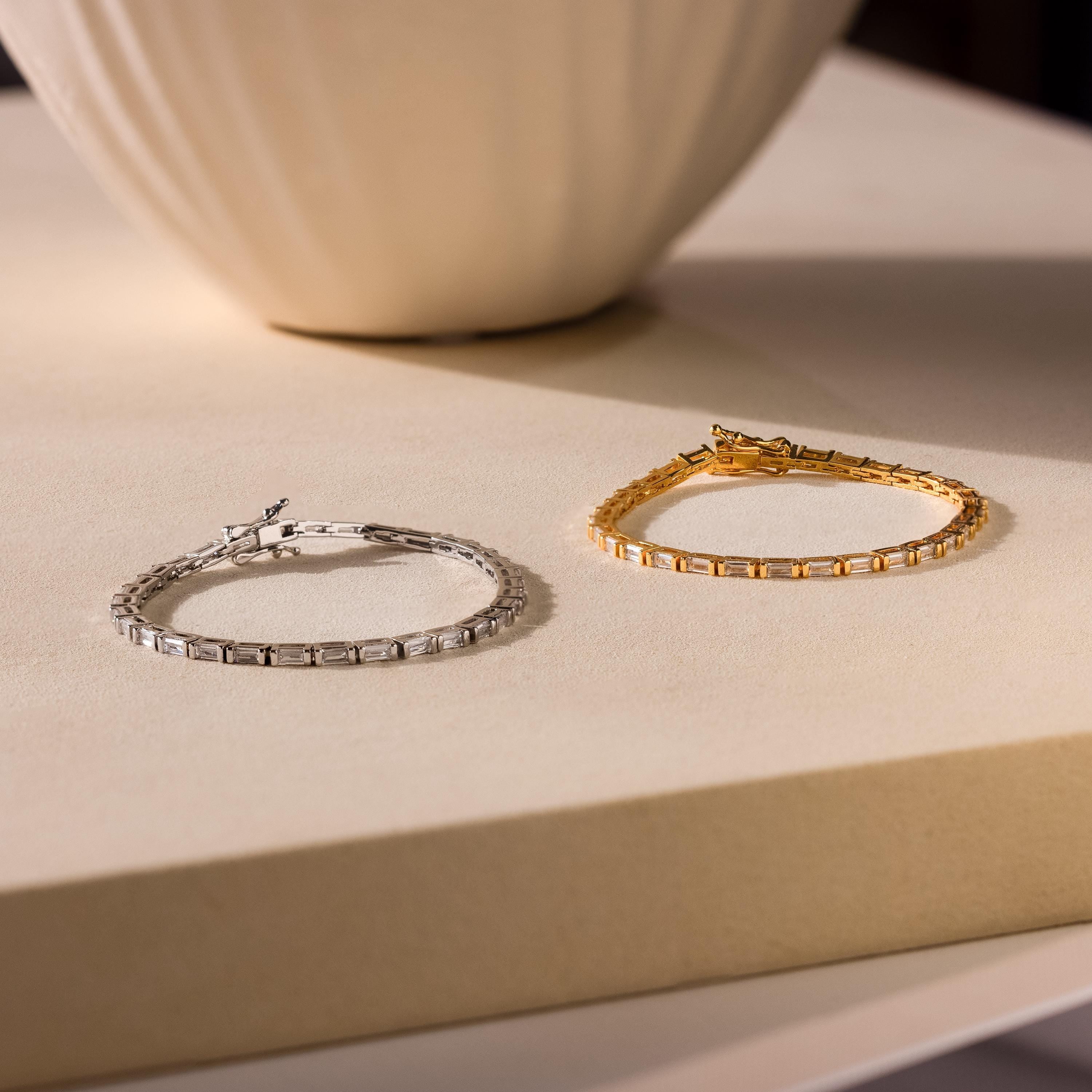 Two Claudia Baguette Tennis Bracelets, in silver and gold, shine on a light beige surface by a vase—an elegant showcase of timeless jewelry.