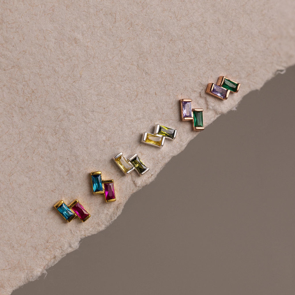 The Duo Baguette Birthstone Studs, vibrant rectangular gemstone earrings ideal for personalized jewelry or birthstone gifts, are displayed diagonally on a textured, torn paper background.