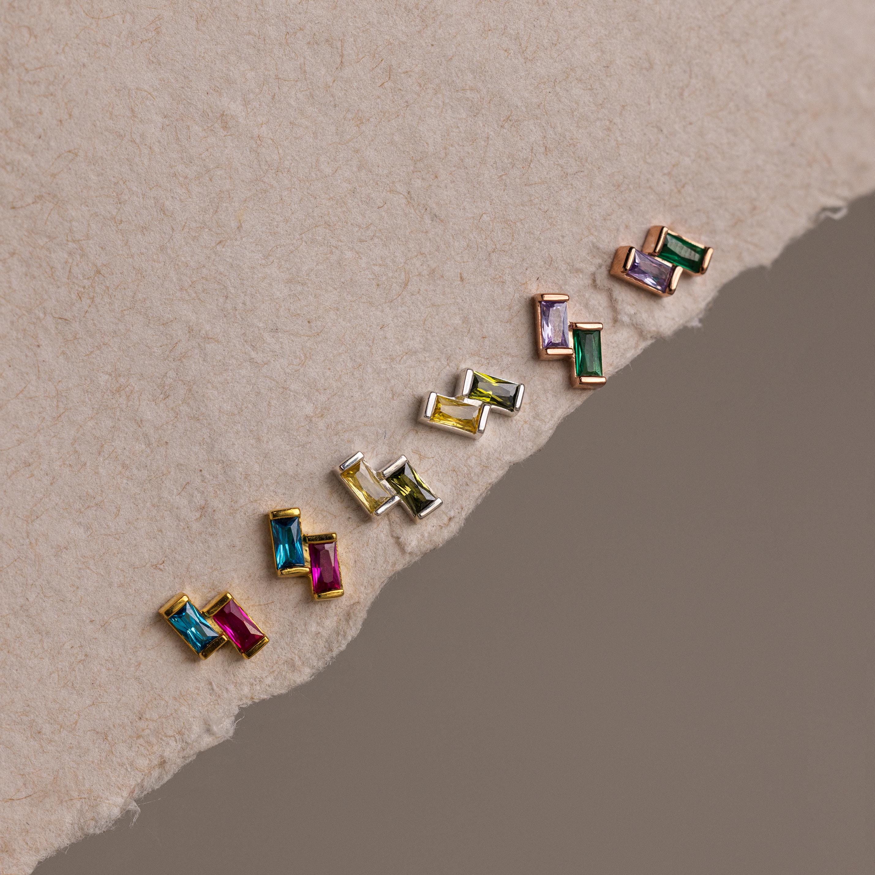 The Duo Baguette Birthstone Studs, vibrant rectangular gemstone earrings ideal for personalized jewelry or birthstone gifts, are displayed diagonally on a textured, torn paper background.
