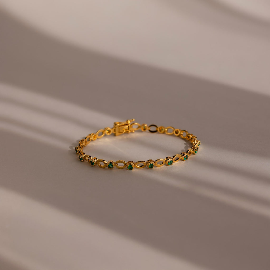 The Emerald Chain Tennis Bracelet features vivid emerald stones set in gold, elegantly displayed on a softly lit surface—an eye-catching birthstone piece ideal for any occasion.