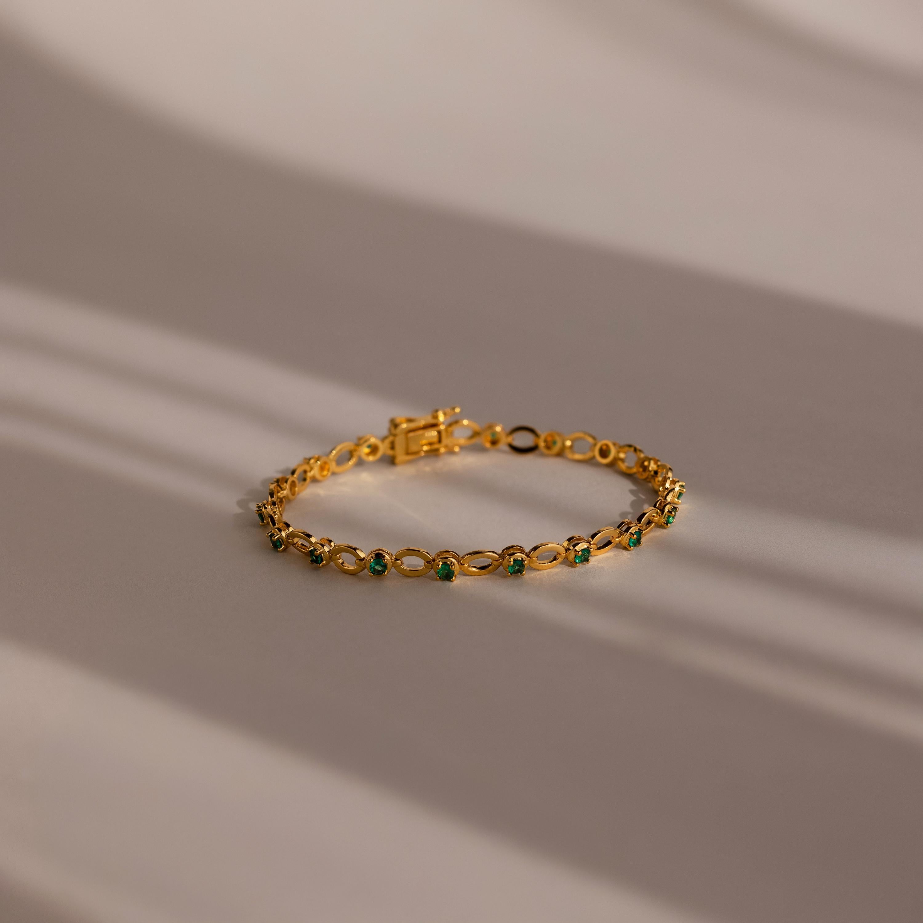 The Emerald Chain Tennis Bracelet features vivid emerald stones set in gold, elegantly displayed on a softly lit surface—an eye-catching birthstone piece ideal for any occasion.
