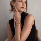 Woman in a sleeveless black top poses with hands near her face, styled with the elegant Pave Paperclip Chain Bracelet featuring gold and pearl accents.
