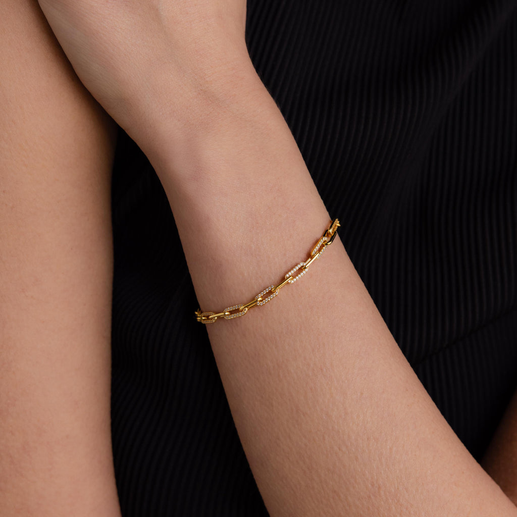 The Pave Paperclip Chain Bracelet elegantly adorns a wrist, showcased against a black textured fabric background.